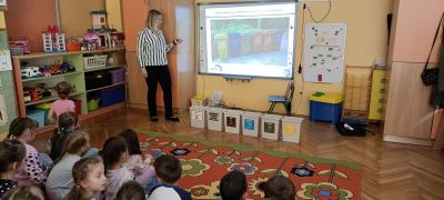 Pani prowadząc zajęcia pokazuje na tablicy kolory koszy do segregowania odpadów. 