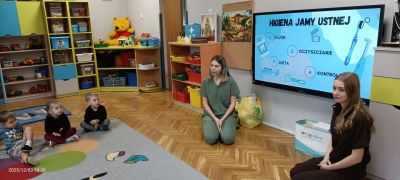 Dzieci siedzą na dywanie i słuchają studentek Stomatologii.